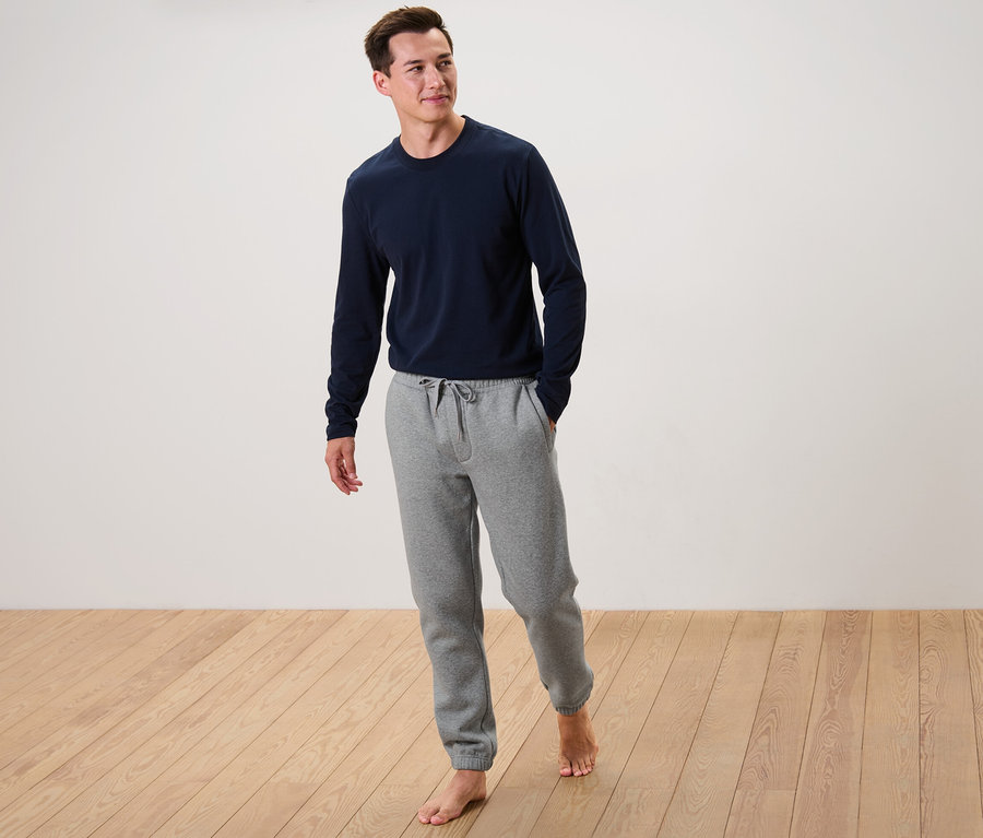 Mann steht barfuß auf Holzboden, trägt ein blaues Langarmshirt und eine graue Sweathose.