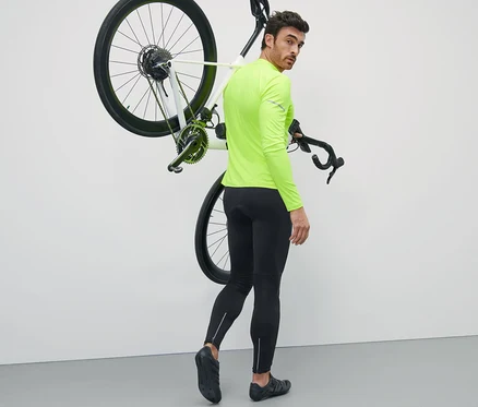 Mann in neongelbem Oberteil trägt ein Fahrrad über der Schulter und trägt eine Fahrradhose.