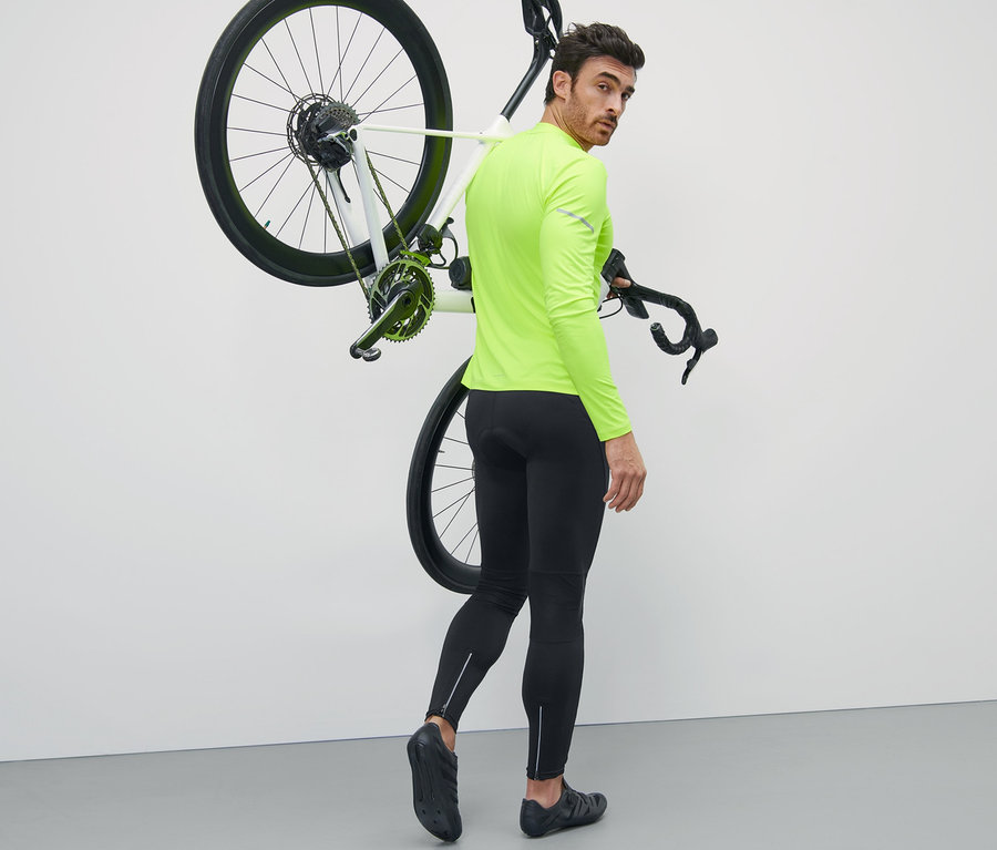 Mann in neongelbem Oberteil trägt ein Fahrrad über der Schulter und trägt eine Fahrradhose.