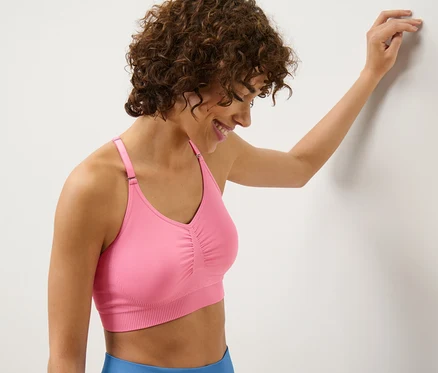 Frau lächelt und lehnt an einer Wand, während sie ein Seamless-Sport-Bustier in Pink trägt.