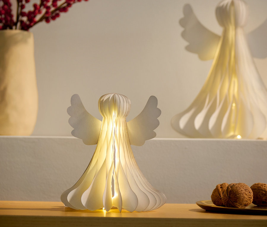 Zwei kleine LED-Papier-Engel, einer auf einem Schrank und der andere auf einem Regal, neben einer Vase mit roten Beeren und einem Teller mit Nüssen.