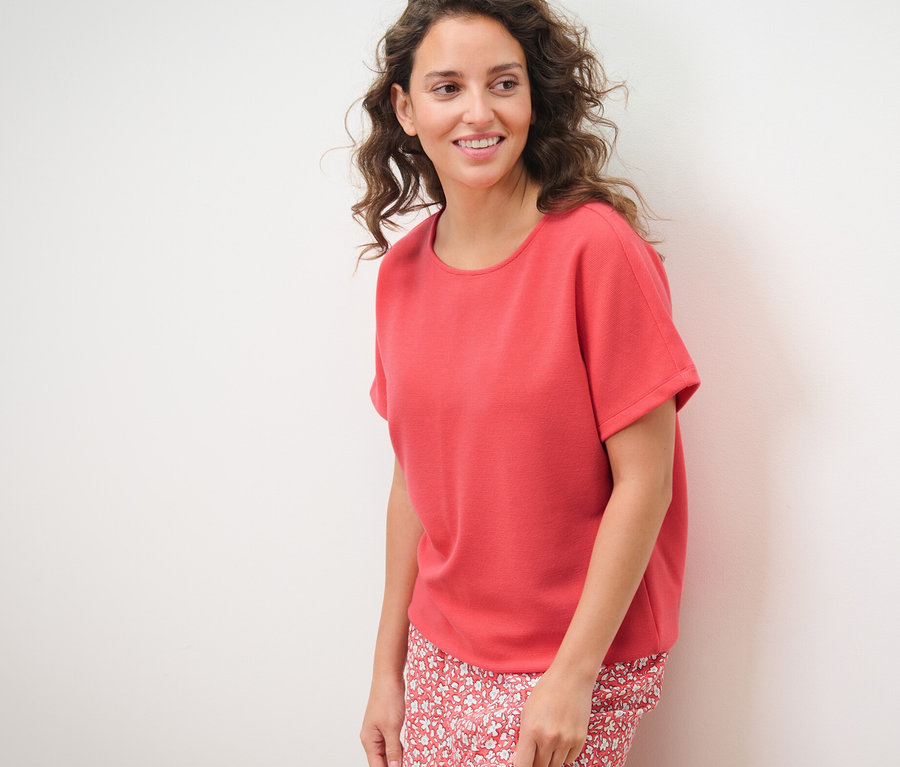 Frau posiert vor einem weißen Hintergrund und trägt ein korallfarbenes Blusenshirt.