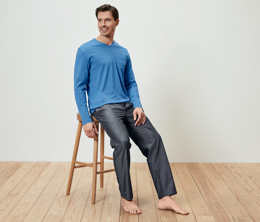 Mann sitzt auf einem Holzhocker und trägt ein blaues Pyjamaoberteil und eine gewebte Hose.