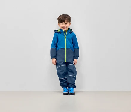 Lächelnder Junge posiert in blauer Jacke mit Kapuze und dunkelblauer Hose mit reflektierenden Streifen.