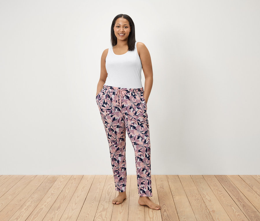 Eine Frau steht mit den Händen in den Taschen einer geblümten Hose. Sie trägt ein weißes Tanktop.
