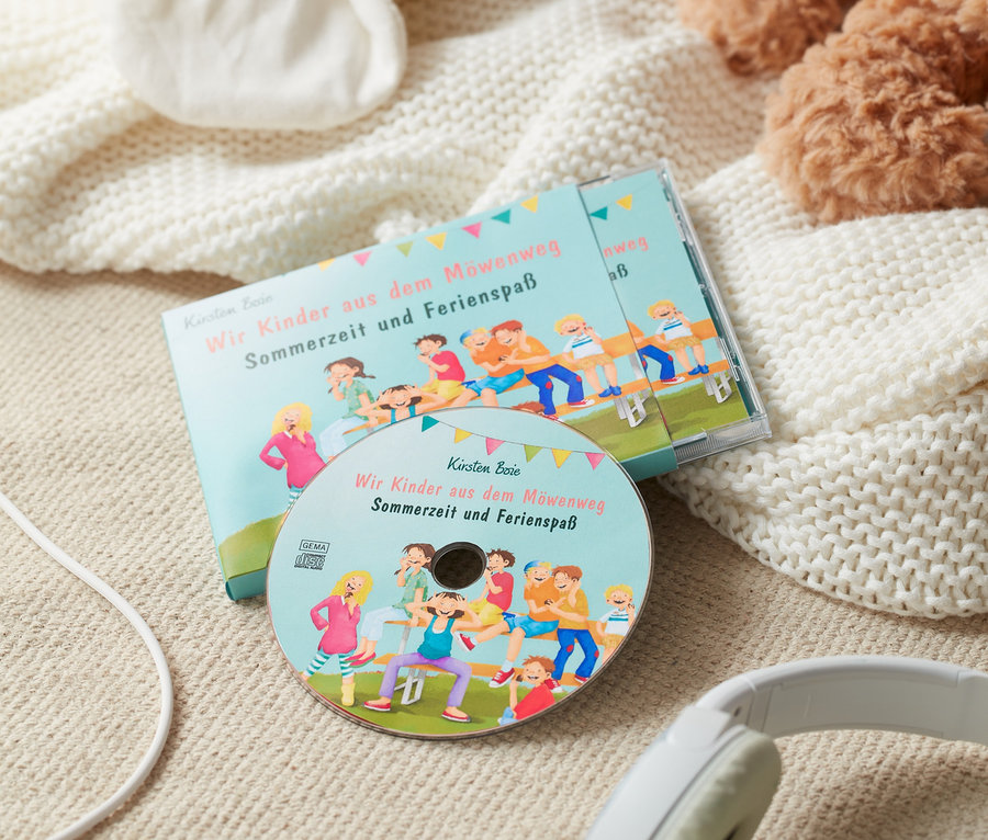 CD-Hörbuch »Wir Kinder aus dem Möwenweg – Sommerzeit und Ferienspaß« liegt mit Kopfhörern auf einer Decke neben einem Stofftier.
