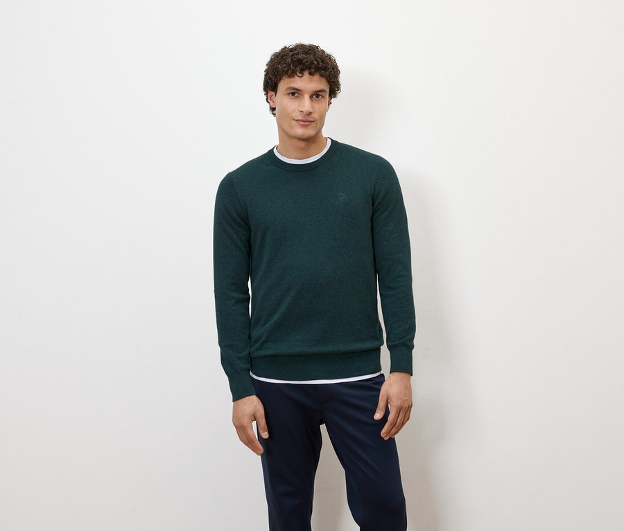 Ein Mann posiert vor einem weißen Hintergrund in einem dunkelgrünen Pullover und dunkelblauen Hosen.