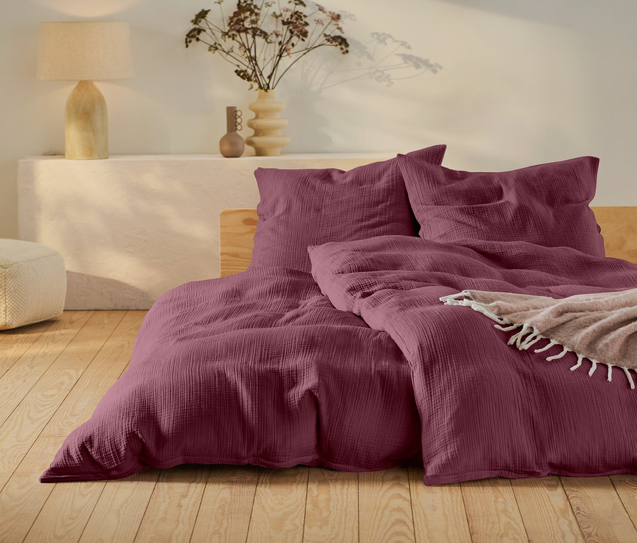 Weinrote Bettwäsche auf einem Bett mit Holzrahmen, ein beiger Pouf und eine Lampe auf einem weißen Tisch.