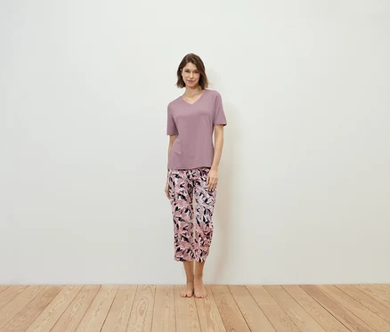 Frau steht und trägt ein violettes T-Shirt und eine geblümte Hose.