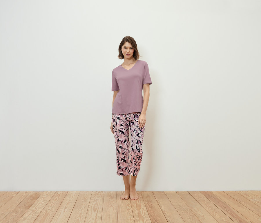 Frau steht und trägt ein violettes T-Shirt und eine geblümte Hose.