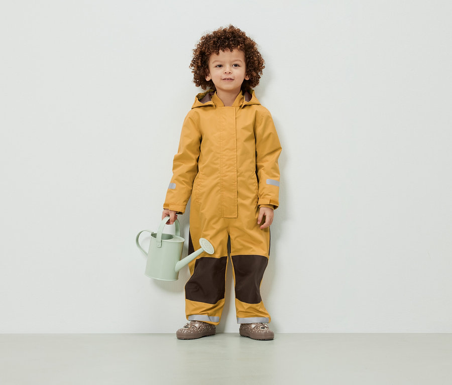 Ein Kind mit lockigem Haar steht in einem gelben Overall und hält eine hellgrüne Gießkanne.