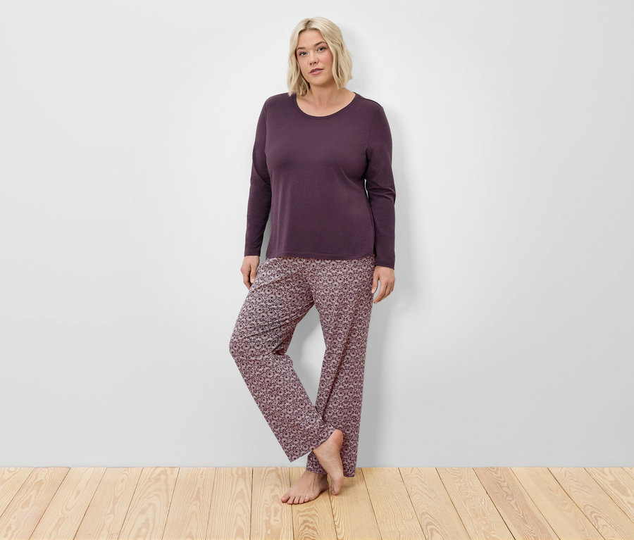 Blonde Frau in weinrotem Langarm-Pyjama und floraler Hose posiert vor einer Wand auf einem Holzboden.