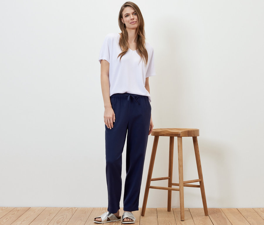 Frau steht neben einem Holzhocker und trägt eine dunkelblaue Leinenhose und ein weißes T-Shirt.