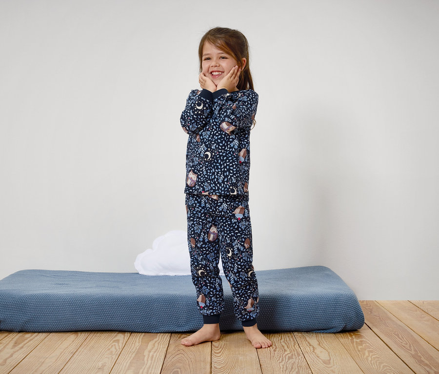 Ein lächelndes Mädchen in einem blauen Pyjama mit Aufdruck steht auf einem Holzboden.