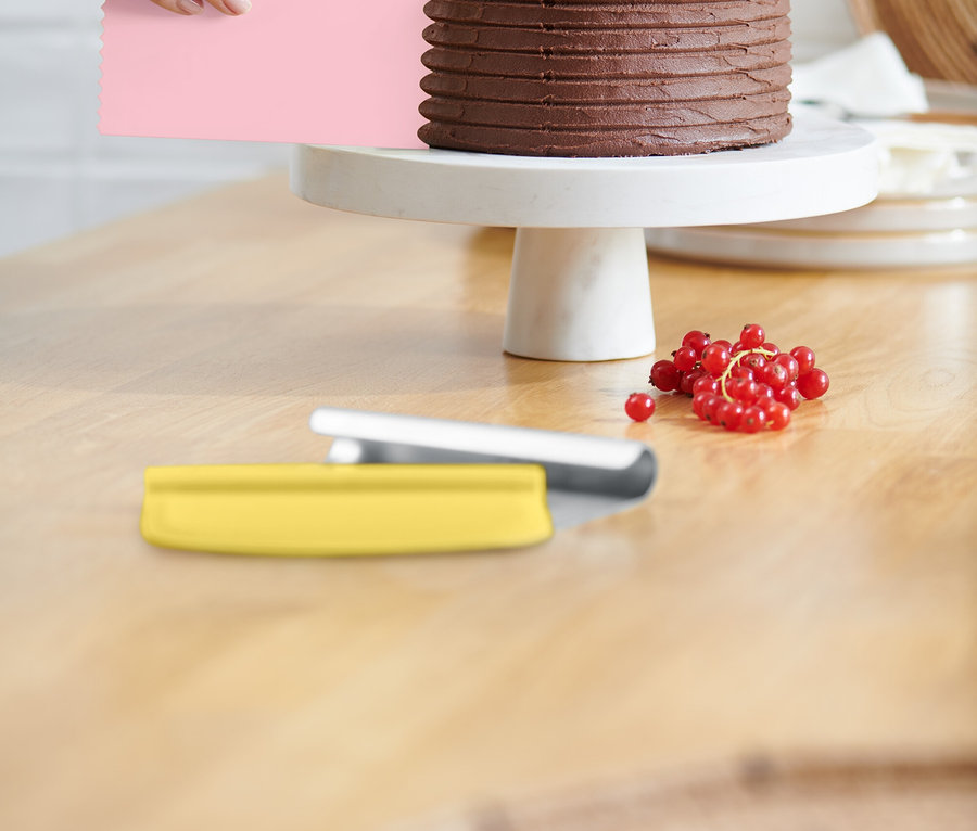 Eine Hand glättet die Glasur einer Schokoladentorte mit einem rosa Teigschaber. Die Torte steht auf einem weißen Tortenständer und ist mit roten Johannisbeeren belegt. Ein gelber Teigschaber liegt auf dem Tisch.