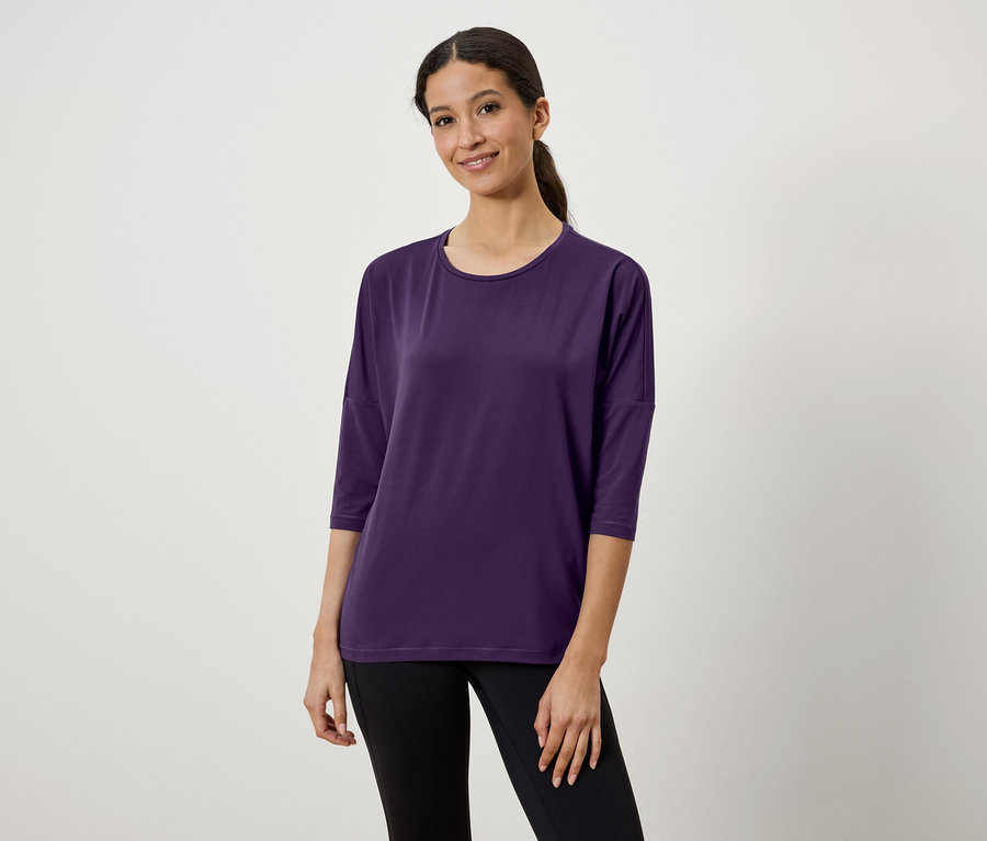 Eine Frau mit braunen Haaren, die zu einem Pferdeschwanz gebunden sind, trägt ein violettes Dreiviertelarm-T-Shirt und schwarze Leggings.