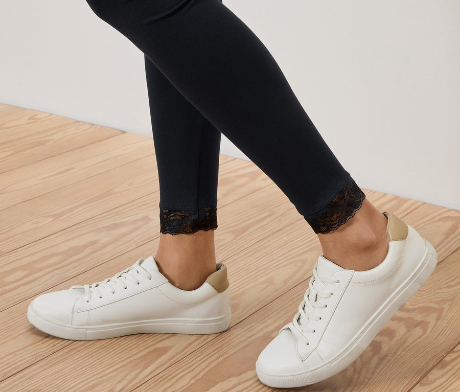 Nahaufnahme von Beinen in schwarzen Leggings mit Spitzenbesatz, kombiniert mit weißen Turnschuhen auf einem Holzboden.