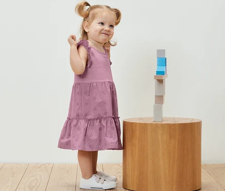 Lächelndes kleines Mädchen im Kinder-Kleid steht neben Holzbausteinen auf einem runden Tisch.
