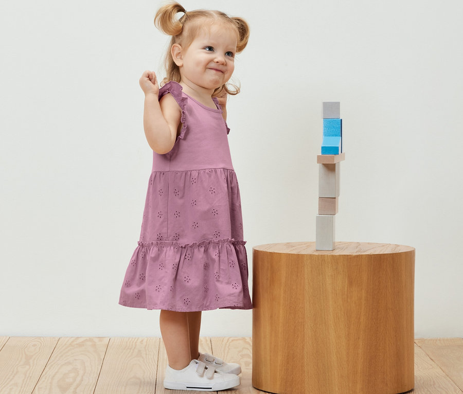 Lächelndes kleines Mädchen im Kinder-Kleid steht neben Holzbausteinen auf einem runden Tisch.