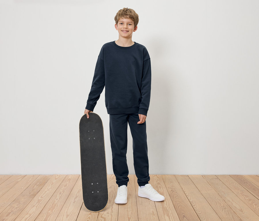 Junge im marineblauen Trainingsanzug hält ein Skateboard.