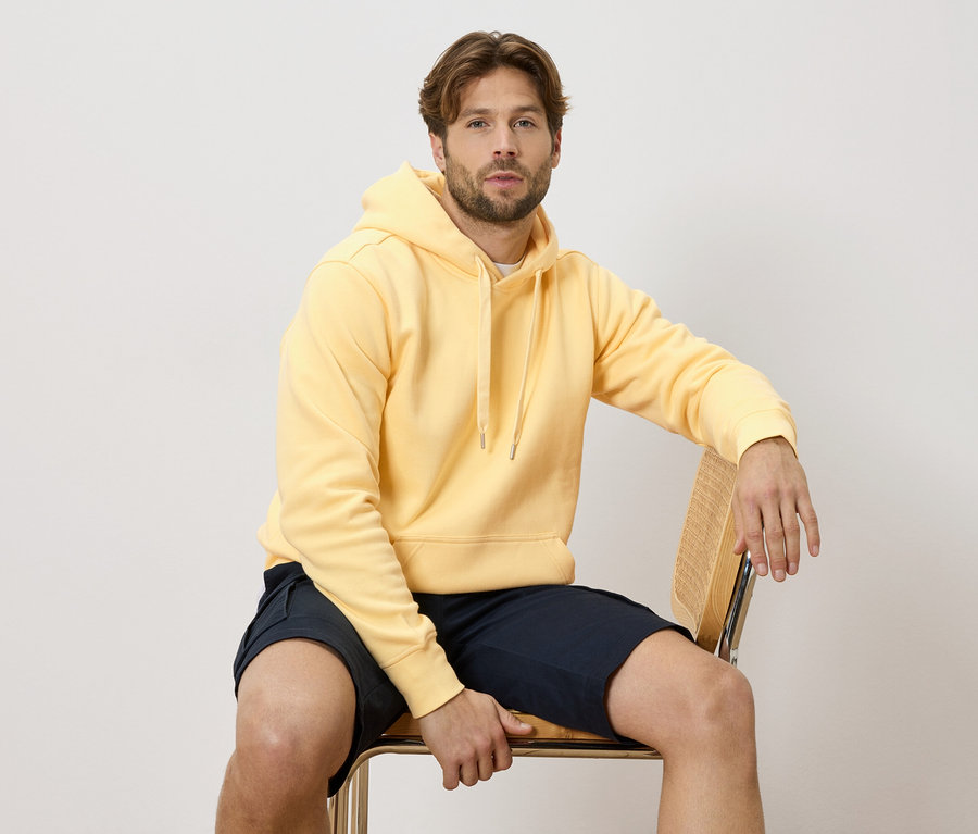 Ein Mann sitzt auf einem Stuhl und trägt einen gelben Hoodie und blaue Shorts.
