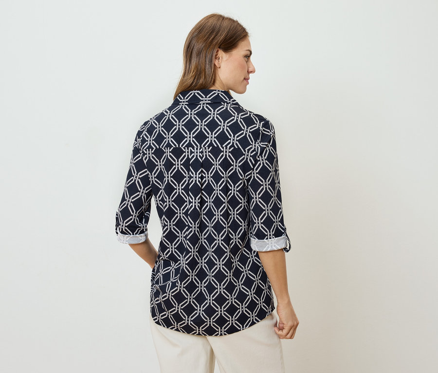 Rückansicht einer Frau mit braunen Haaren, die eine marineblaue, langärmlige Bluse mit weißem geometrischem Muster und beige Hosen trägt.