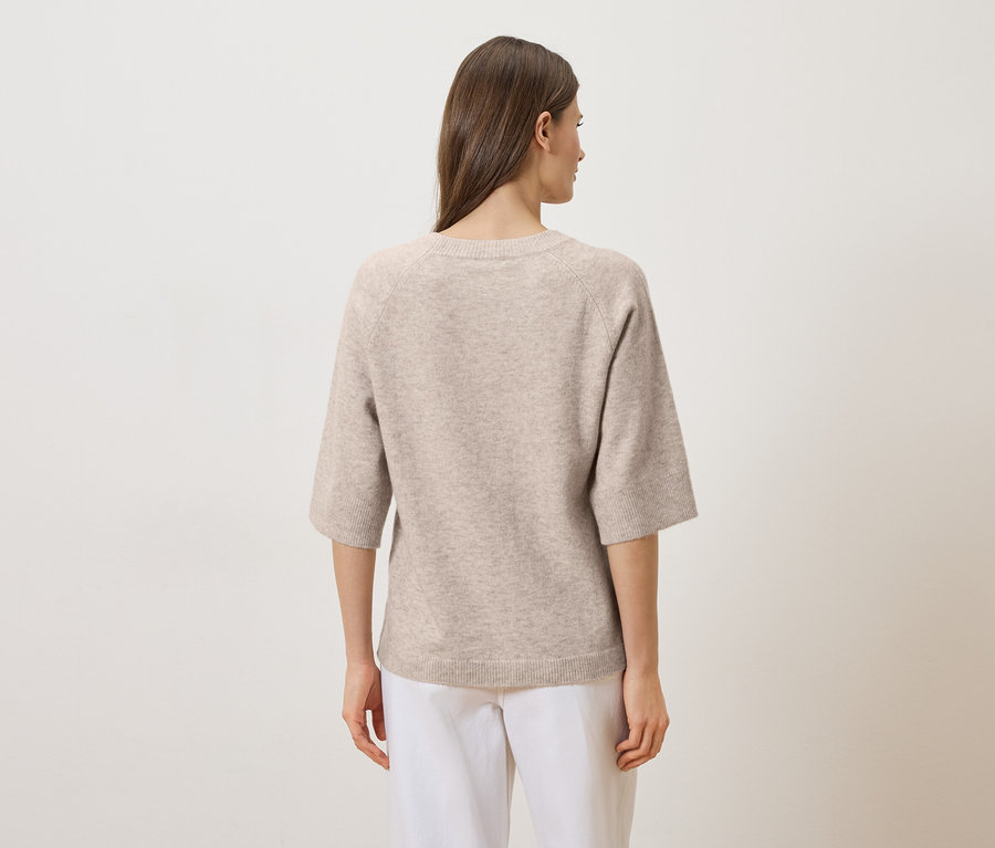 Eine Frau mit langen braunen Haaren trägt einen beigen Pullover mit Dreiviertelärmeln und weiße Hosen, von hinten gesehen.