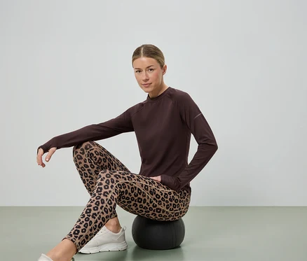 Frau sitzt auf einem schwarzen Ball, trägt ein braunes Langarmshirt, Leggings mit Leopardenmuster und weiße Turnschuhe.