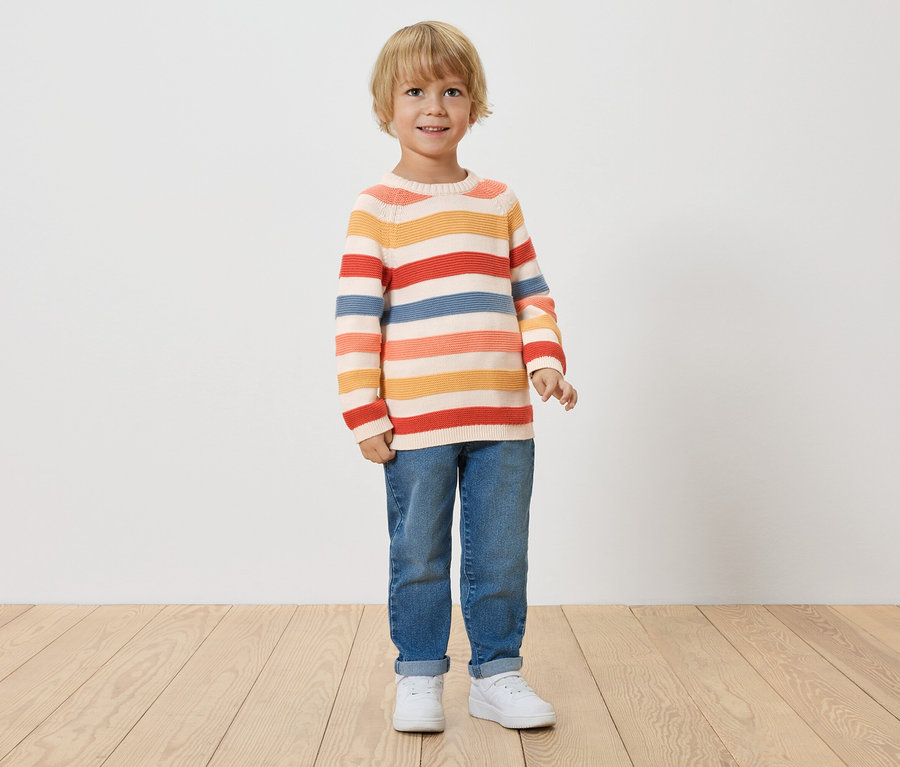 Ein kleiner Junge mit blonden Haaren steht vor einer weißen Wand und trägt einen gestreiften Pullover, Jeans und weiße Turnschuhe.