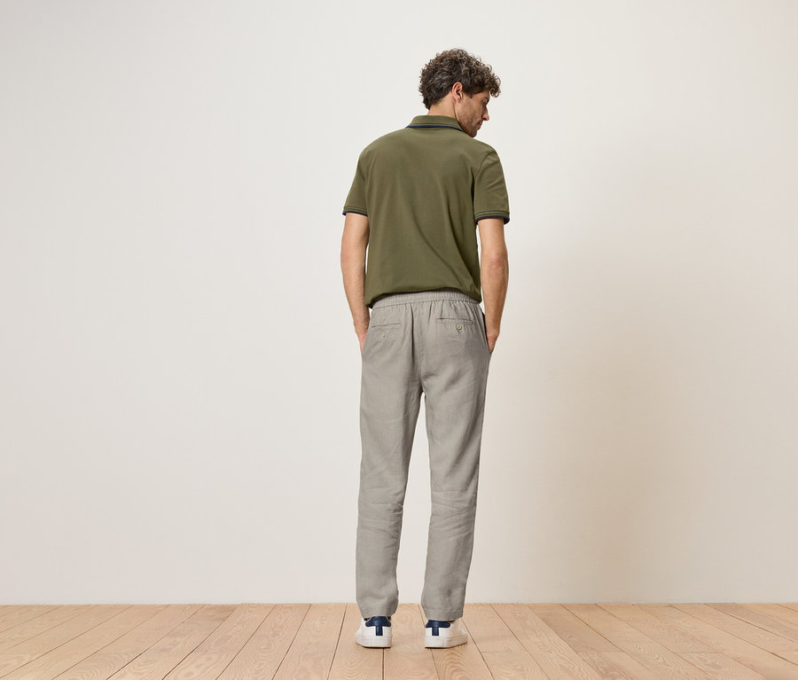Ein Mann mit lockigem Haar steht mit dem Rücken zur Kamera, trägt ein olivgrünes Poloshirt, graue Leinenhosen und weiße Schuhe mit blauen Details.