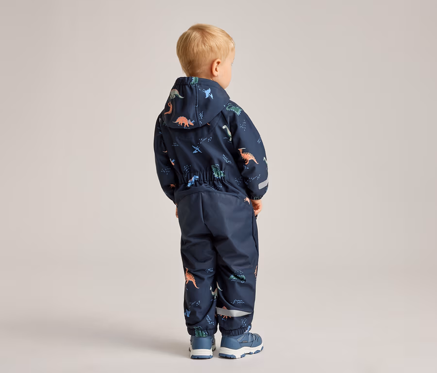 Ein Kind steht mit dem Rücken zur Kamera und trägt einen marineblauen Overall mit Dinosaurier-Muster und blaue Schuhe.