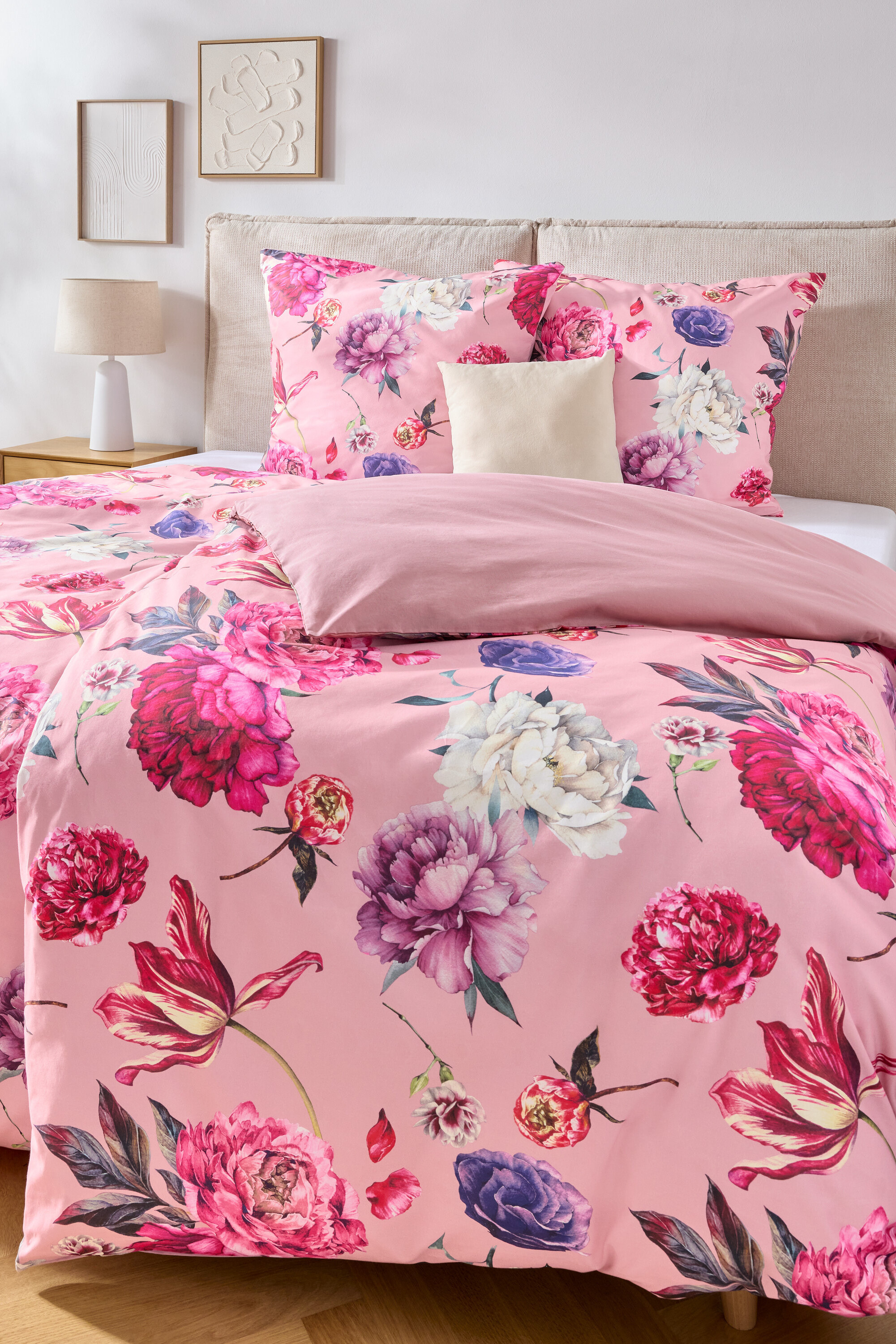 Nahaufnahme eines Bettes mit rosa Bettwäsche mit Blumenmuster, einem beigen Kopfteil und zwei gerahmten Bildern an der Wand.