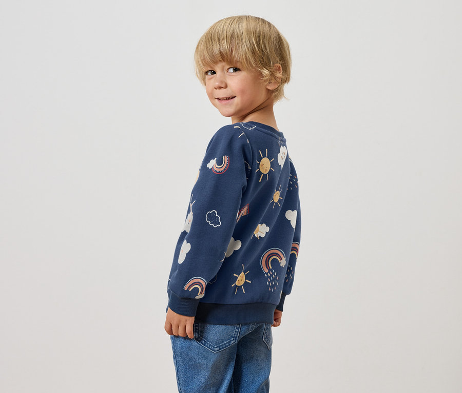 Ein Junge mit blonden Haaren blickt über seine Schulter und trägt ein blaues Sweatshirt mit Regenbogen-, Wolken- und Sonnenmotiven.
