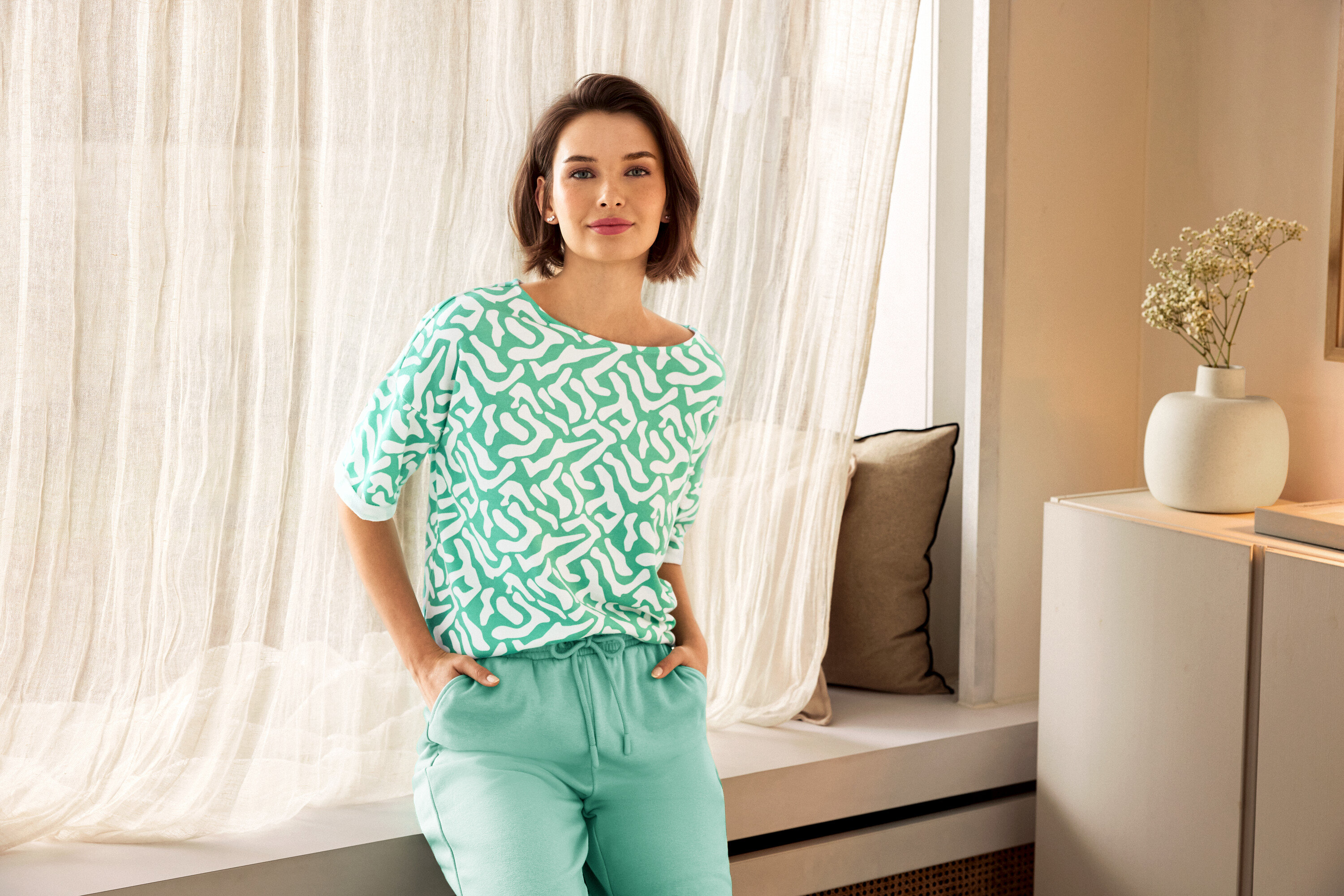 Frau mit grüner Hose und gemustertem Oberteil posiert neben einem Fenster. Im Hintergrund sind 925 Silber Ohrstecker Zirkoniabogen und eine Vase zu sehen.