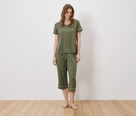Eine Frau steht barfuß auf einem Holzboden und trägt einen olivgrünen Pyjama mit weißen Akzenten.