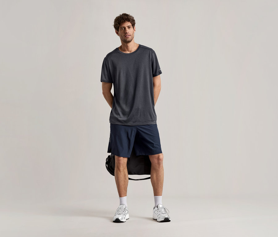 Mann steht mit Sportshirt, anthrazitfarben, Shorts und Turnschuhen vor hellem Hintergrund.