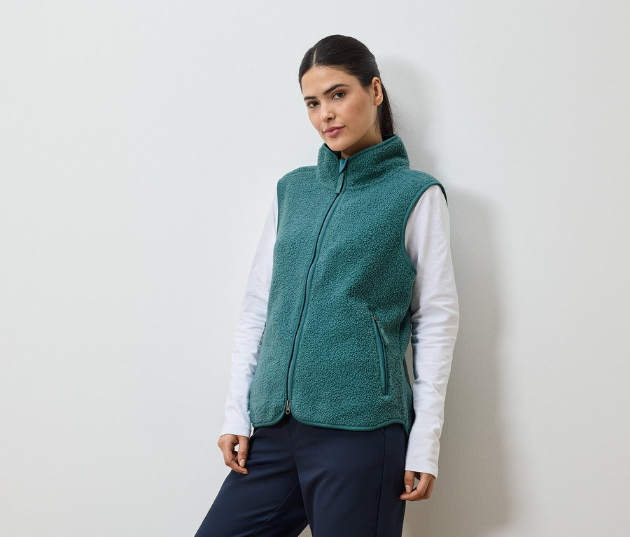 Frau mit Pferdeschwanz, die eine grüne Fleeceweste über einem weißen Langarmshirt und einer marineblauen Hose trägt.