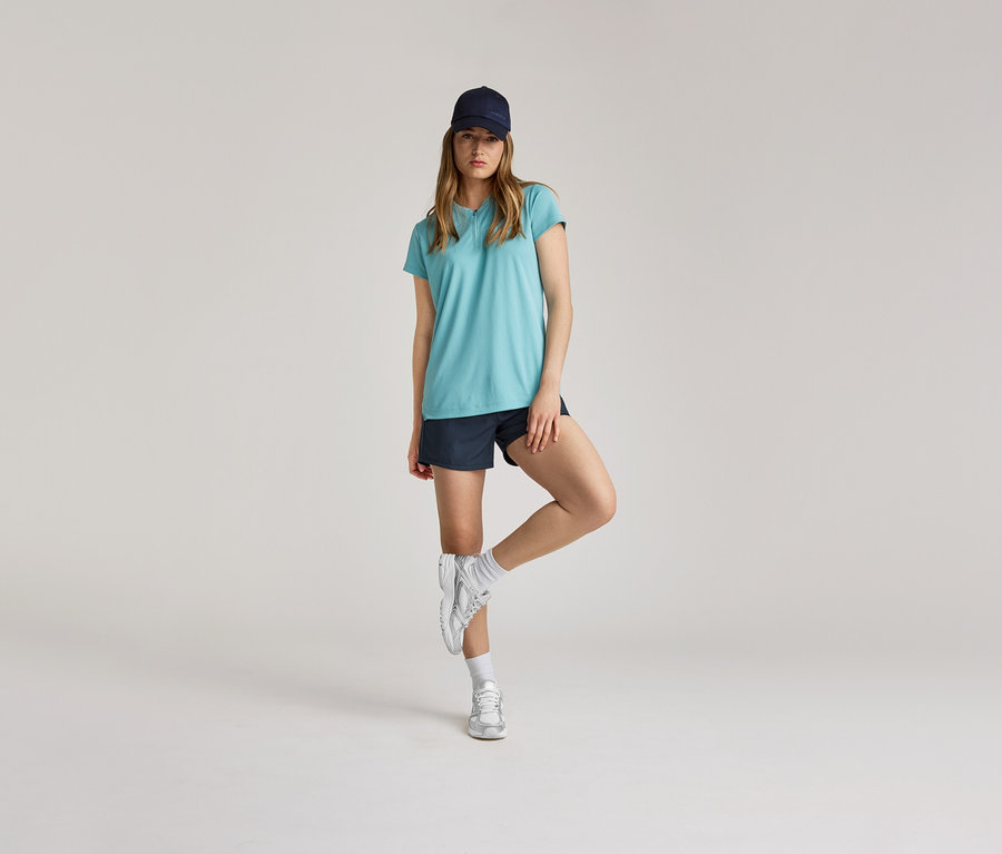Frau in blauem T-Shirt, Shorts und Mütze steht auf einem Bein vor hellem Hintergrund.