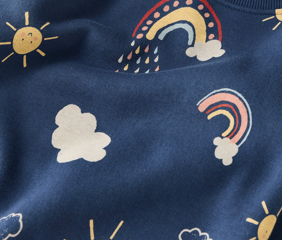 Nahaufnahme eines dunkelblauen Stoffes mit sich wiederholenden Mustern aus gelben Sonnen, weißen Wolken und bunten Regenbögen.