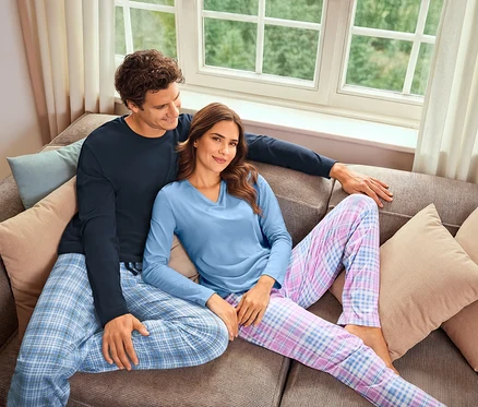 Ein Mann und eine Frau sitzen auf einem Sofa. Der Mann trägt einen marineblauen Pyjama und die Frau einen hellblauen Pyjama mit Karomuster.