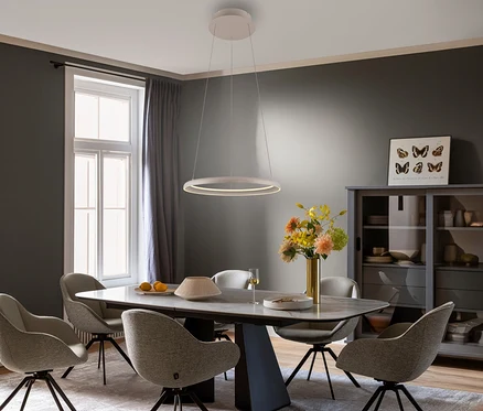 Ein modernes Esszimmer mit einem rechteckigen Tisch, sechs hellen Stühlen und einer weißen, ringförmigen SCHÖNER WOHNEN-Kollektion Pendelleuchte »Quillo«, sandbrise darüber. Der Raum hat dunkelgraue Wände, ein großes Fenster mit grauen Vorhängen und eine graue Vitrine.