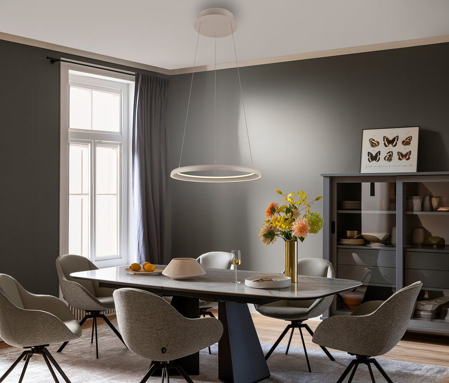 Ein modernes Esszimmer mit einem rechteckigen Tisch, sechs hellen Stühlen und einer weißen, ringförmigen SCHÖNER WOHNEN-Kollektion Pendelleuchte »Quillo«, sandbrise darüber. Der Raum hat dunkelgraue Wände, ein großes Fenster mit grauen Vorhängen und eine graue Vitrine.