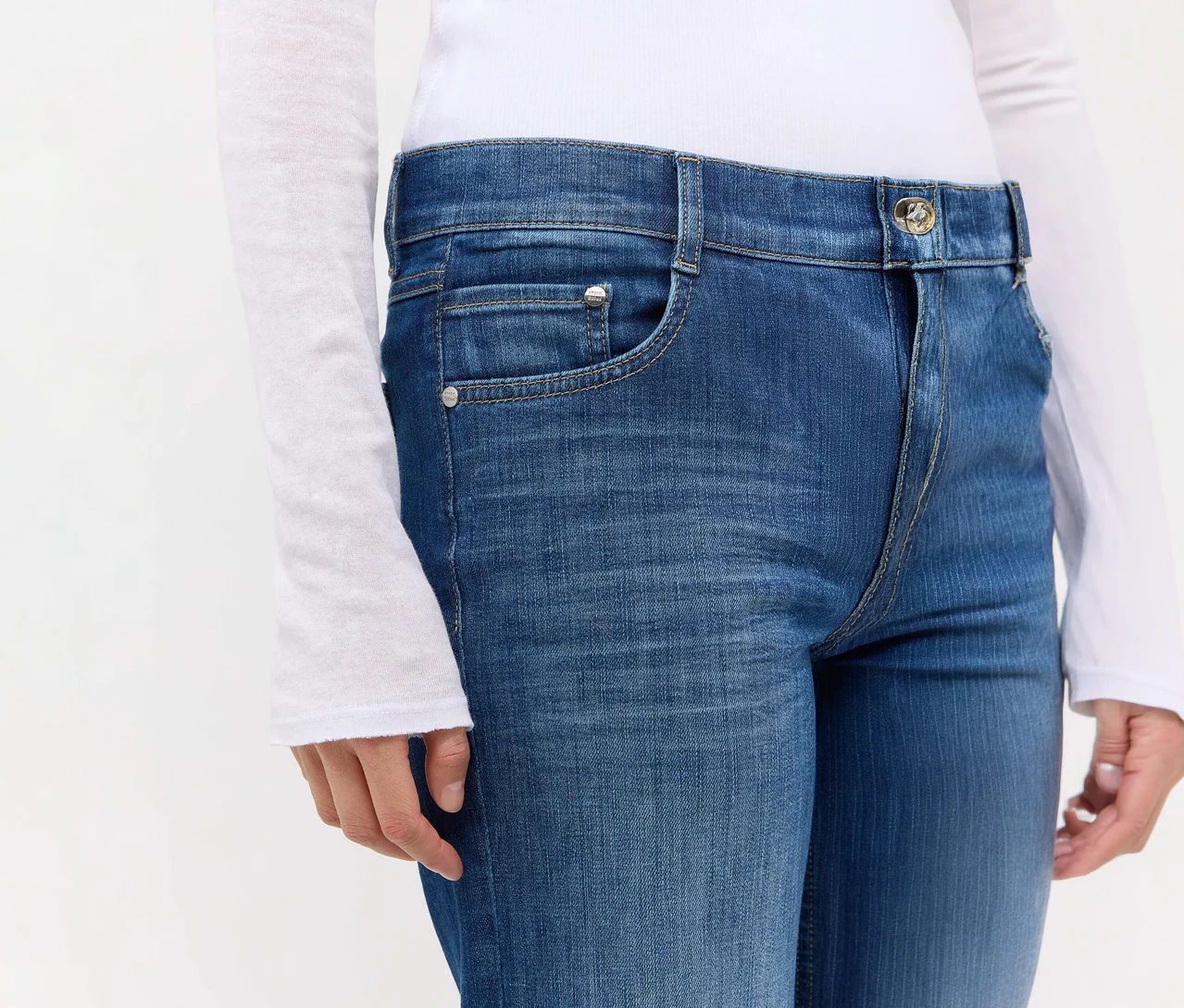 Person trägt mittelblaue Jeans mit Waschung, Vordertaschen und Knopfverschluss, kombiniert mit einem weißen Langarmshirt.