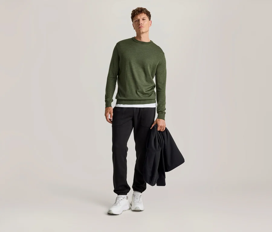 Mann steht, trägt einen grünen Langarmpullover über einem weißen T-Shirt, schwarze Hose und weiße Turnschuhe. Er hält eine schwarze Jacke in der Hand.