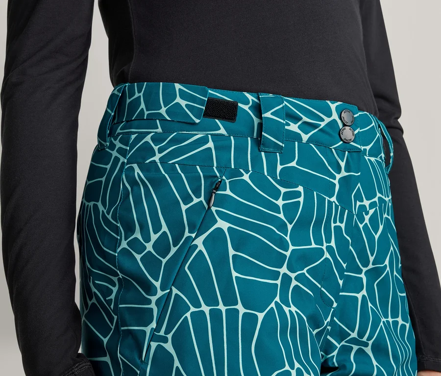 Nahaufnahme einer Person in einem schwarzen Langarmshirt und einer tealfarbenen Hose mit hellblauem, abstraktem Muster. Die Hose hat einen Bund mit Knöpfen und eine Reißverschlusstasche.