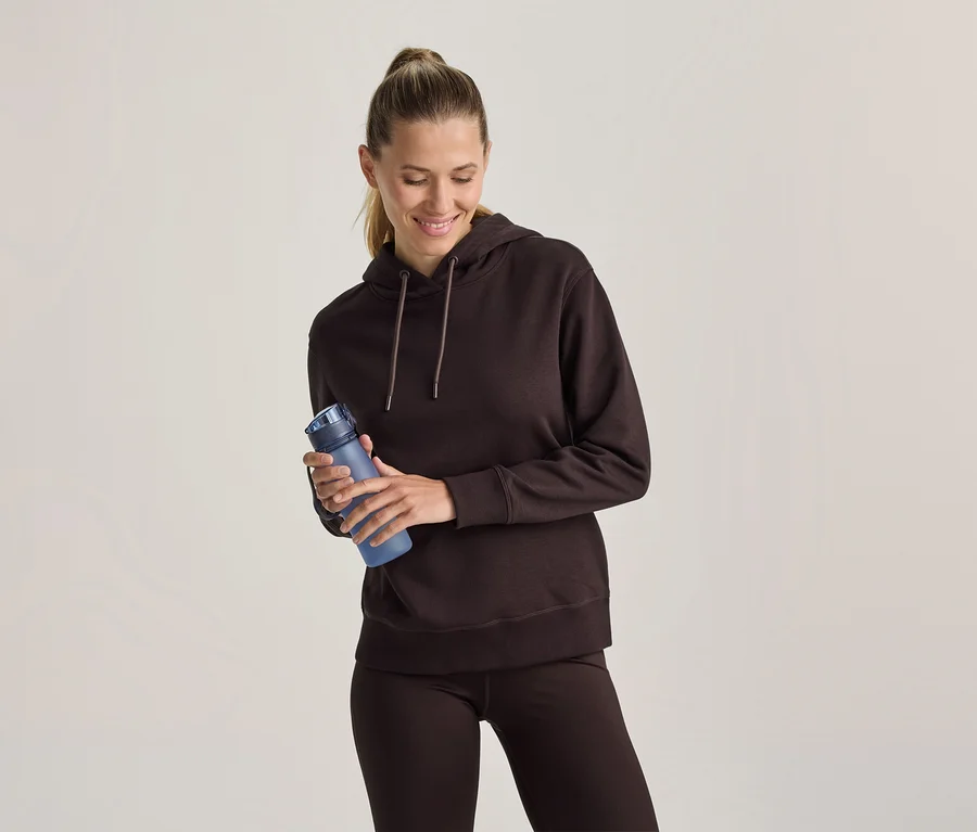 Frau in einem braunen Hoodie und braunen Leggings hält eine blaue Trinkflasche, lächelt und blickt nach unten.