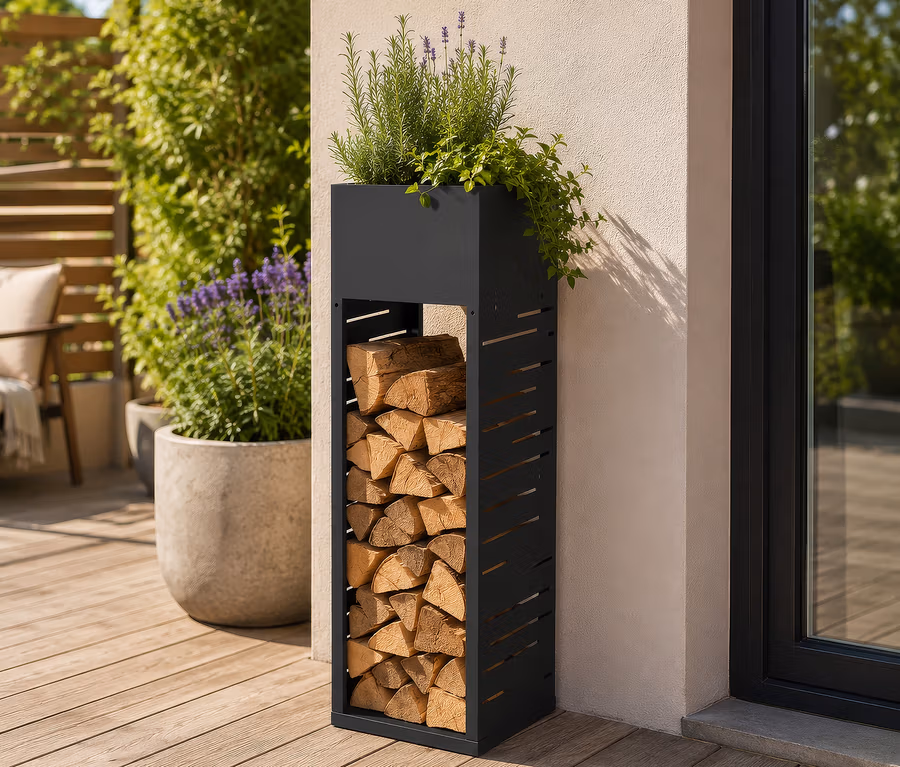 Ein schwarzes Metall-Holzregal mit gestapeltem Brennholz, obenauf ein Pflanzgefäß mit Grünpflanzen und Lavendel, steht auf einer Holzterrasse neben einer hellen Wand und einer Glastür.