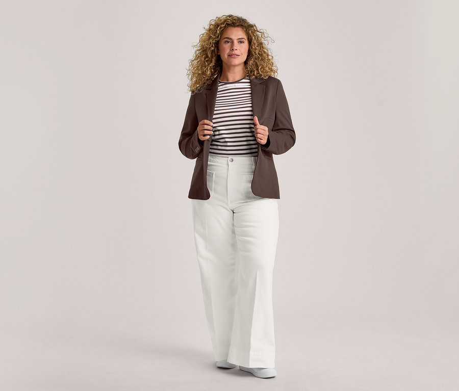Eine Frau mit lockigem Haar steht frontal, hält die Revers ihres offenen braunen Blazers. Sie trägt ein schwarz-weiß gestreiftes T-Shirt, eine weiße weite Hose und helle flache Schuhe.