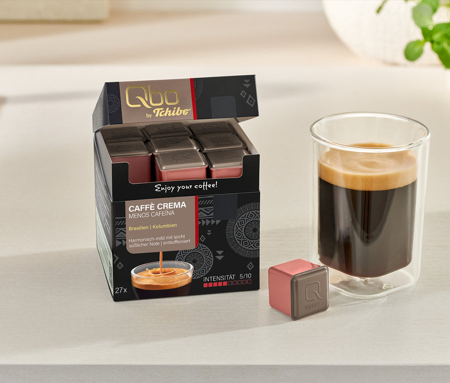 Eine geöffnete Packung Qbo Caffè MENOS CAFEÍNA mit 27 Kapseln, eine Kapsel und ein Glas Kaffee stehen auf einer hellen Oberfläche.