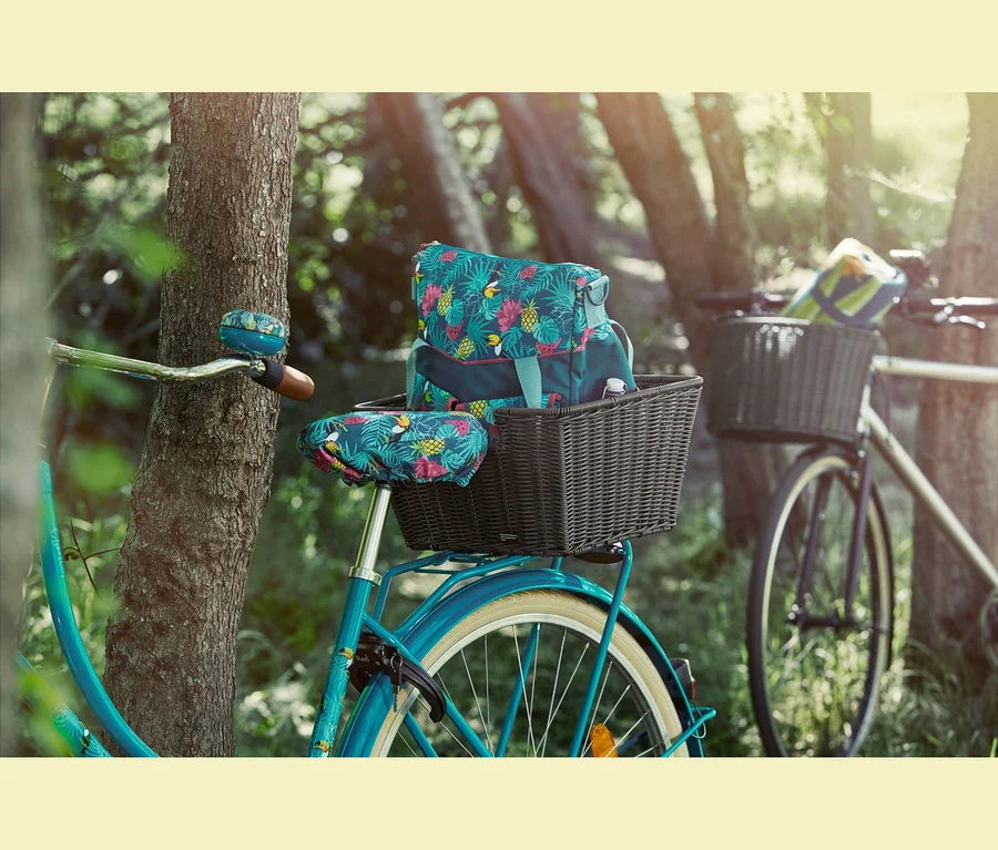 Ein blaues Fahrrad lehnt an einem Baum. Auf dem Gepäckträger befindet sich ein Fahrradkorb mit einer Rad- und Umhängetasche darin. Am Lenker ist eine Fahrradklingel und auf dem Sattel ein Fahrrad-Sattelbezug.